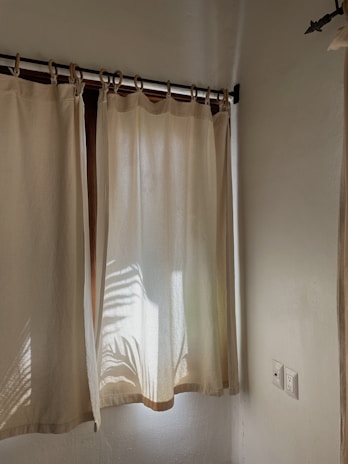 Light beige curtains hang on a wooden rod, with sunlight casting the shadow of a palm leaf onto the fabric. The room's walls are painted in a light cream color, and there is an electrical outlet visible near the bottom right corner.