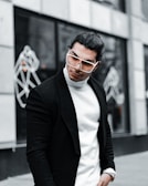 A stylish person with long hair is wearing oversized sunglasses, a white turtleneck sweater, and a black coat. The background features abstract line art on a building facade, giving a modern and urban feel.