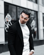 A stylish person with long hair is wearing oversized sunglasses, a white turtleneck sweater, and a black coat. The background features abstract line art on a building facade, giving a modern and urban feel.