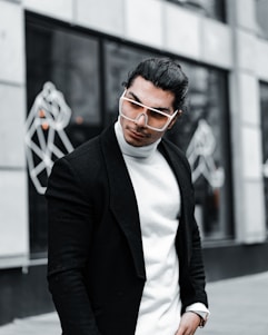 A stylish person with long hair is wearing oversized sunglasses, a white turtleneck sweater, and a black coat. The background features abstract line art on a building facade, giving a modern and urban feel.