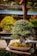 A carefully sculpted bonsai tree sits in a rectangular ceramic pot, its roots covered with a layer of moss. The tree is full of dense green foliage with small leaves and is placed on a wooden table. In the background, other trees and garden elements can be seen out of focus.
