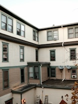 An external view of a multi-story residential building with cream-colored siding and dark green trim around the windows. The building features multiple windows, some with air conditioning units. Two satellite dishes are mounted on the wall. The roof shows parts of an adjacent structure with a flat surface. A ladder and some outdoor objects are located near the wall, surrounded by a few bare tree branches.