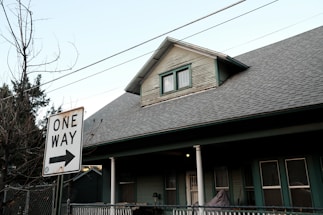 A residential house with a pitched roof and a smaller upper window. There is a 'One Way' street sign in the foreground pointing to the right. Bare trees and utility wires stretch across the sky, while the house features a porch with white columns and green siding.