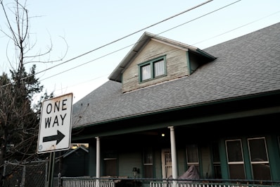 A residential house with a pitched roof and a smaller upper window. There is a 'One Way' street sign in the foreground pointing to the right. Bare trees and utility wires stretch across the sky, while the house features a porch with white columns and green siding.