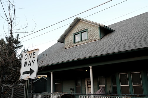 A residential house with a pitched roof and a smaller upper window. There is a 'One Way' street sign in the foreground pointing to the right. Bare trees and utility wires stretch across the sky, while the house features a porch with white columns and green siding.
