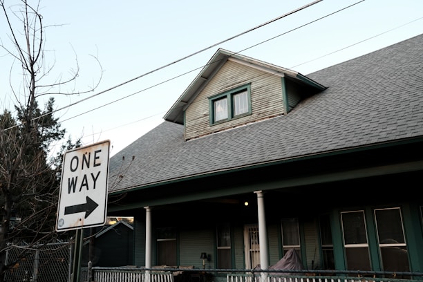 A residential house with a pitched roof and a smaller upper window. There is a 'One Way' street sign in the foreground pointing to the right. Bare trees and utility wires stretch across the sky, while the house features a porch with white columns and green siding.