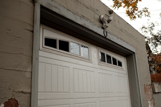 A large white garage door set in a beige concrete structure. Above the garage door, there are windows with three of the small panes being blacked out. A security light is mounted above the door. There are some visible signs of wear and paint coming off on the building and the surrounding autumn foliage is visible at the top right corner of the image.