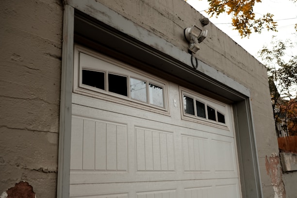 A large white garage door set in a beige concrete structure. Above the garage door, there are windows with three of the small panes being blacked out. A security light is mounted above the door. There are some visible signs of wear and paint coming off on the building and the surrounding autumn foliage is visible at the top right corner of the image.