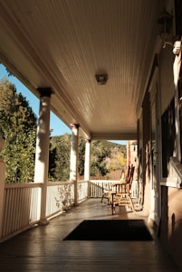 A cozy wooden back porch with potted herbs, a rocking chair, and sunlight filtering through leafy trees.