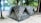 Two camouflaged tents are set up on a sandy ground surrounded by trees. The tents are positioned side by side in a natural forest setting, close to a beach or water body. The area is shaded by numerous trees, creating a peaceful and secluded campground atmosphere.