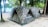Two camouflaged tents are set up on a sandy ground surrounded by trees. The tents are positioned side by side in a natural forest setting, close to a beach or water body. The area is shaded by numerous trees, creating a peaceful and secluded campground atmosphere.