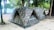 Two camouflaged tents are set up on a sandy ground surrounded by trees. The tents are positioned side by side in a natural forest setting, close to a beach or water body. The area is shaded by numerous trees, creating a peaceful and secluded campground atmosphere.
