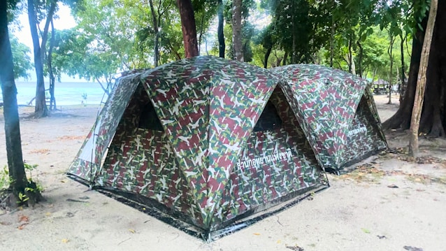 Two camouflaged tents are set up on a sandy ground surrounded by trees. The tents are positioned side by side in a natural forest setting, close to a beach or water body. The area is shaded by numerous trees, creating a peaceful and secluded campground atmosphere.