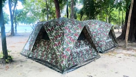 Two camouflaged tents are set up on a sandy ground surrounded by trees. The tents are positioned side by side in a natural forest setting, close to a beach or water body. The area is shaded by numerous trees, creating a peaceful and secluded campground atmosphere.