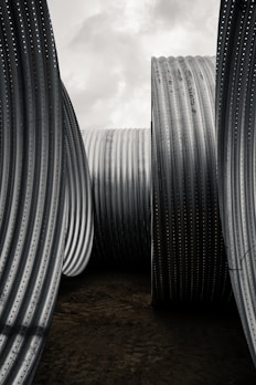 Large, textured metal pipes stand vertically under a cloudy sky, creating an industrial and monumental feel. The corrugated surfaces and the sheer size of the pipes emphasize their sturdiness and functional design.
