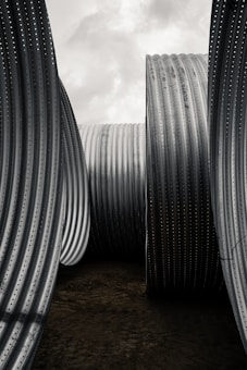 Large, textured metal pipes stand vertically under a cloudy sky, creating an industrial and monumental feel. The corrugated surfaces and the sheer size of the pipes emphasize their sturdiness and functional design.