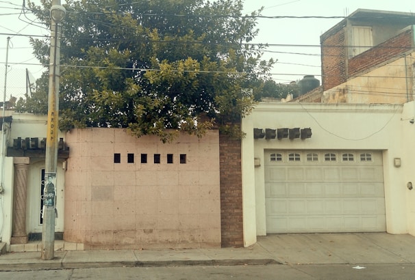 A street view featuring a residential facade with a large garage door and a small section of wall with square openings. A lush tree is visible above the wall, providing a touch of greenery. An adjacent lamp post has a poster attached, and various overhead cables are strung across the scene.