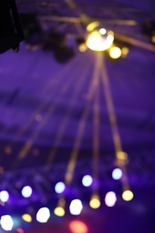 Bright, colorful stage lights create rays and lens flares against a blurred background, conveying a lively and dynamic atmosphere.