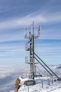 A tall metallic tower equipped with various antennas and instruments is situated on a snowy mountain peak. The scene overlooks a vast expanse of clouds with hints of distant mountains under a clear blue sky.