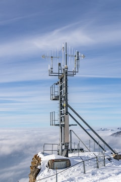A tall metallic tower equipped with various antennas and instruments is situated on a snowy mountain peak. The scene overlooks a vast expanse of clouds with hints of distant mountains under a clear blue sky.