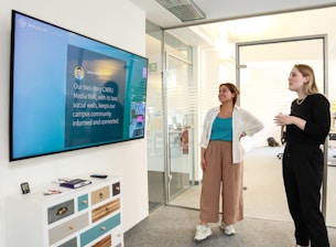 Two people are standing in an office setting, looking at a large wall-mounted screen displaying a message about a media wall. One person is holding a drink, and the environment includes a chest of drawers, glass walls, and overhead lights.