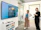 Two people are standing in an office setting, looking at a large wall-mounted screen displaying a message about a media wall. One person is holding a drink, and the environment includes a chest of drawers, glass walls, and overhead lights.