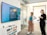 Two people are standing in an office setting, looking at a large wall-mounted screen displaying a message about a media wall. One person is holding a drink, and the environment includes a chest of drawers, glass walls, and overhead lights.