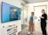 Two people are standing in an office setting, looking at a large wall-mounted screen displaying a message about a media wall. One person is holding a drink, and the environment includes a chest of drawers, glass walls, and overhead lights.