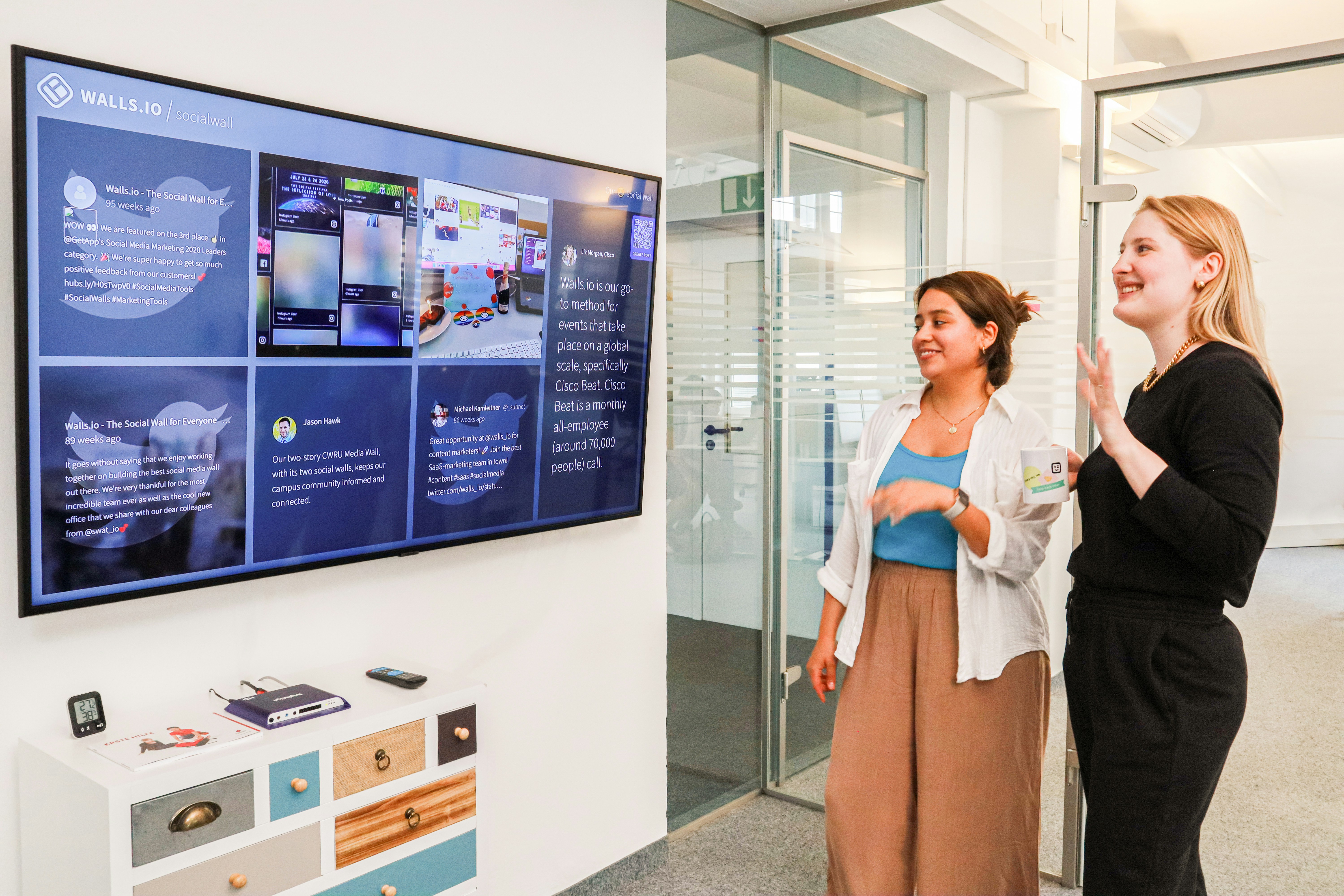 Two women stand in an office environment, looking at a large digital display on the wall showcasing a social media feed. One woman is smiling and gesturing with her hand. The area is modern and well-lit, with a small white cabinet featuring multicolored drawers in the foreground. Clear glass doors and partitions define the space, indicating a professional setting.