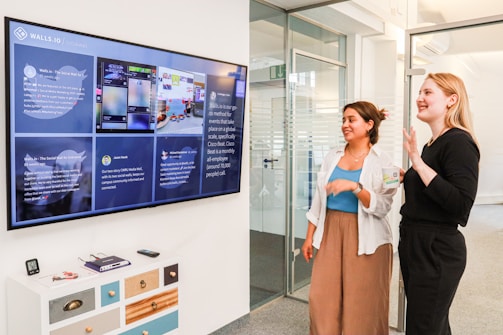 Two women stand in an office environment, looking at a large digital display on the wall showcasing a social media feed. One woman is smiling and gesturing with her hand. The area is modern and well-lit, with a small white cabinet featuring multicolored drawers in the foreground. Clear glass doors and partitions define the space, indicating a professional setting.