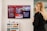 A woman with blonde hair is standing in an office setting, looking at a large screen display mounted on the wall. The screen displays a social media wall with multiple posts arranged neatly. The background reveals a modern office environment with a sideboard featuring colorful drawers and various electronic devices placed on top.