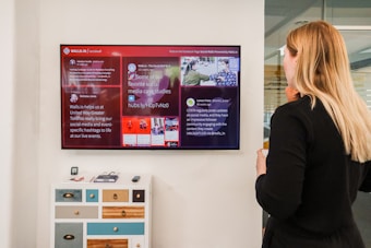A woman with blonde hair is standing in an office setting, looking at a large screen display mounted on the wall. The screen displays a social media wall with multiple posts arranged neatly. The background reveals a modern office environment with a sideboard featuring colorful drawers and various electronic devices placed on top.