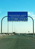 A blue road sign indicating directions to Al Khafji Port in the Kingdom of Saudi Arabia. It provides separate lanes for passengers and cargo trucks. Cars are seen traveling on the highway beneath a clear blue sky.