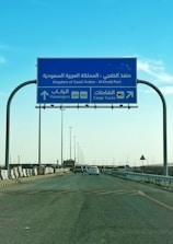 A blue road sign indicating directions to Al Khafji Port in the Kingdom of Saudi Arabia. It provides separate lanes for passengers and cargo trucks. Cars are seen traveling on the highway beneath a clear blue sky.
