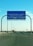 A blue road sign indicating directions to Al Khafji Port in the Kingdom of Saudi Arabia. It provides separate lanes for passengers and cargo trucks. Cars are seen traveling on the highway beneath a clear blue sky.