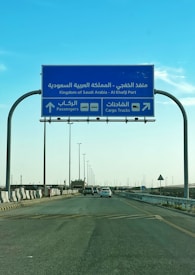 A blue road sign indicating directions to Al Khafji Port in the Kingdom of Saudi Arabia. It provides separate lanes for passengers and cargo trucks. Cars are seen traveling on the highway beneath a clear blue sky.