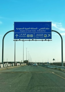 A blue road sign indicating directions to Al Khafji Port in the Kingdom of Saudi Arabia. It provides separate lanes for passengers and cargo trucks. Cars are seen traveling on the highway beneath a clear blue sky.