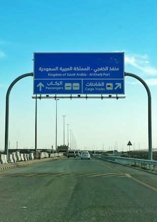 A blue road sign indicating directions to Al Khafji Port in the Kingdom of Saudi Arabia. It provides separate lanes for passengers and cargo trucks. Cars are seen traveling on the highway beneath a clear blue sky.