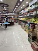A well-organized grocery store aisle with a variety of items displayed on shelves. The right side is stocked with different kinds of fruits like apples and mangoes, while the left side and back wall hold packaged goods and beverages. The ceiling features bright, evenly spaced recessed lights and ceiling fans. Two people are seen standing at a counter, possibly engaging in a transaction.