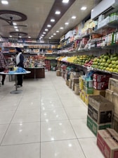 A well-organized grocery store aisle with a variety of items displayed on shelves. The right side is stocked with different kinds of fruits like apples and mangoes, while the left side and back wall hold packaged goods and beverages. The ceiling features bright, evenly spaced recessed lights and ceiling fans. Two people are seen standing at a counter, possibly engaging in a transaction.