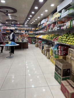 A well-organized grocery store aisle with a variety of items displayed on shelves. The right side is stocked with different kinds of fruits like apples and mangoes, while the left side and back wall hold packaged goods and beverages. The ceiling features bright, evenly spaced recessed lights and ceiling fans. Two people are seen standing at a counter, possibly engaging in a transaction.