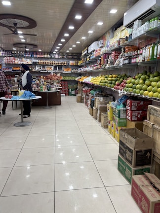 A well-organized grocery store aisle with a variety of items displayed on shelves. The right side is stocked with different kinds of fruits like apples and mangoes, while the left side and back wall hold packaged goods and beverages. The ceiling features bright, evenly spaced recessed lights and ceiling fans. Two people are seen standing at a counter, possibly engaging in a transaction.