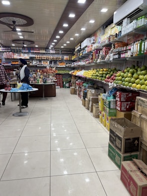 A well-organized grocery store aisle with a variety of items displayed on shelves. The right side is stocked with different kinds of fruits like apples and mangoes, while the left side and back wall hold packaged goods and beverages. The ceiling features bright, evenly spaced recessed lights and ceiling fans. Two people are seen standing at a counter, possibly engaging in a transaction.