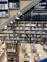 A modern bookstore featuring multiple shelves filled with various books and magazines. The layout includes a large wall of books with clean, organized rows. Below, there are additional tables showcasing more books. The setting has a contemporary design with bright lighting, and a section of the wall has signage for a family restaurant.