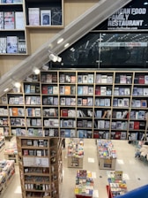 A modern bookstore featuring multiple shelves filled with various books and magazines. The layout includes a large wall of books with clean, organized rows. Below, there are additional tables showcasing more books. The setting has a contemporary design with bright lighting, and a section of the wall has signage for a family restaurant.
