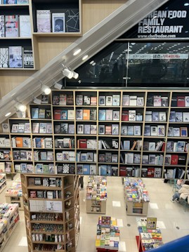 A modern bookstore featuring multiple shelves filled with various books and magazines. The layout includes a large wall of books with clean, organized rows. Below, there are additional tables showcasing more books. The setting has a contemporary design with bright lighting, and a section of the wall has signage for a family restaurant.