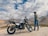 A man stands beside a motorcycle in a rugged, mountainous landscape. The sky is partly cloudy, and the terrain consists of rocky ground and distant mountains. The motorbike is positioned prominently, with visible branding. The man, wearing casual outdoor attire, gazes into the distance.