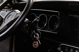 A close-up view of a car interior focusing on the dashboard and steering wheel. The dashboard features classic analog dials, and there is a set of keys hanging with a tag that reads 'MINI'. The steering wheel is positioned in the foreground and shows part of a logo.