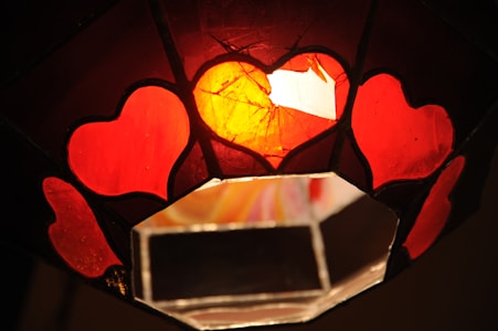 A stained glass artwork featuring heart shapes with vivid red and orange tones. The hearts are outlined with black leading, and the center glows warmly against the darker surrounding glass.