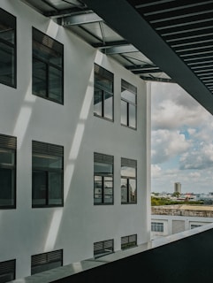 A modern building facade with several large windows under a slatted metal roof. Sunlight casts shadows across the white exterior wall. In the background, there is a view of a cityscape with a tall building, trees, and a cloudy sky.